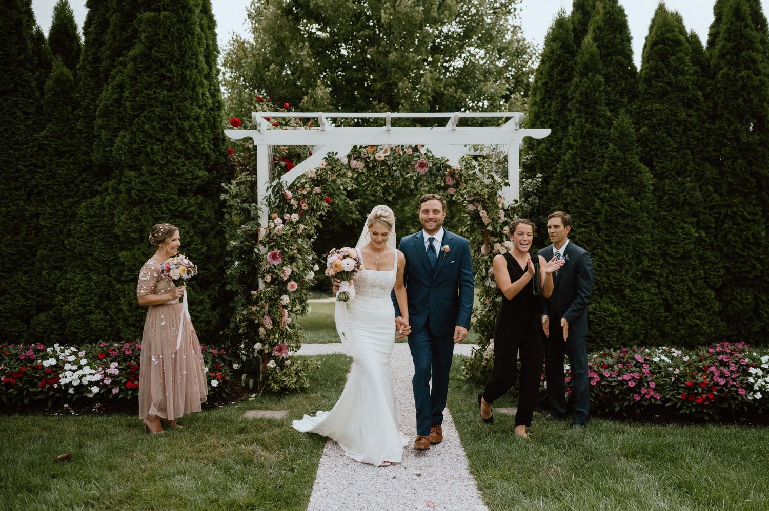 Newlyweds exit their garden ceremony under a white pergola decorated with pink flowers.