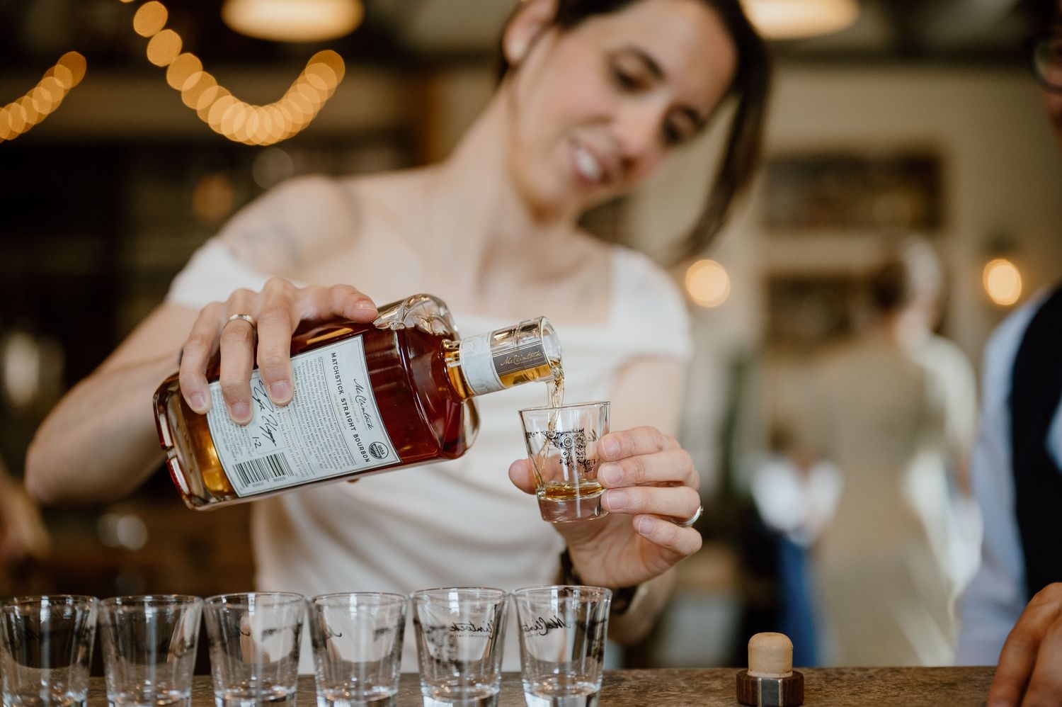 Person in white pouring spirits into glasses at a distillery bar.