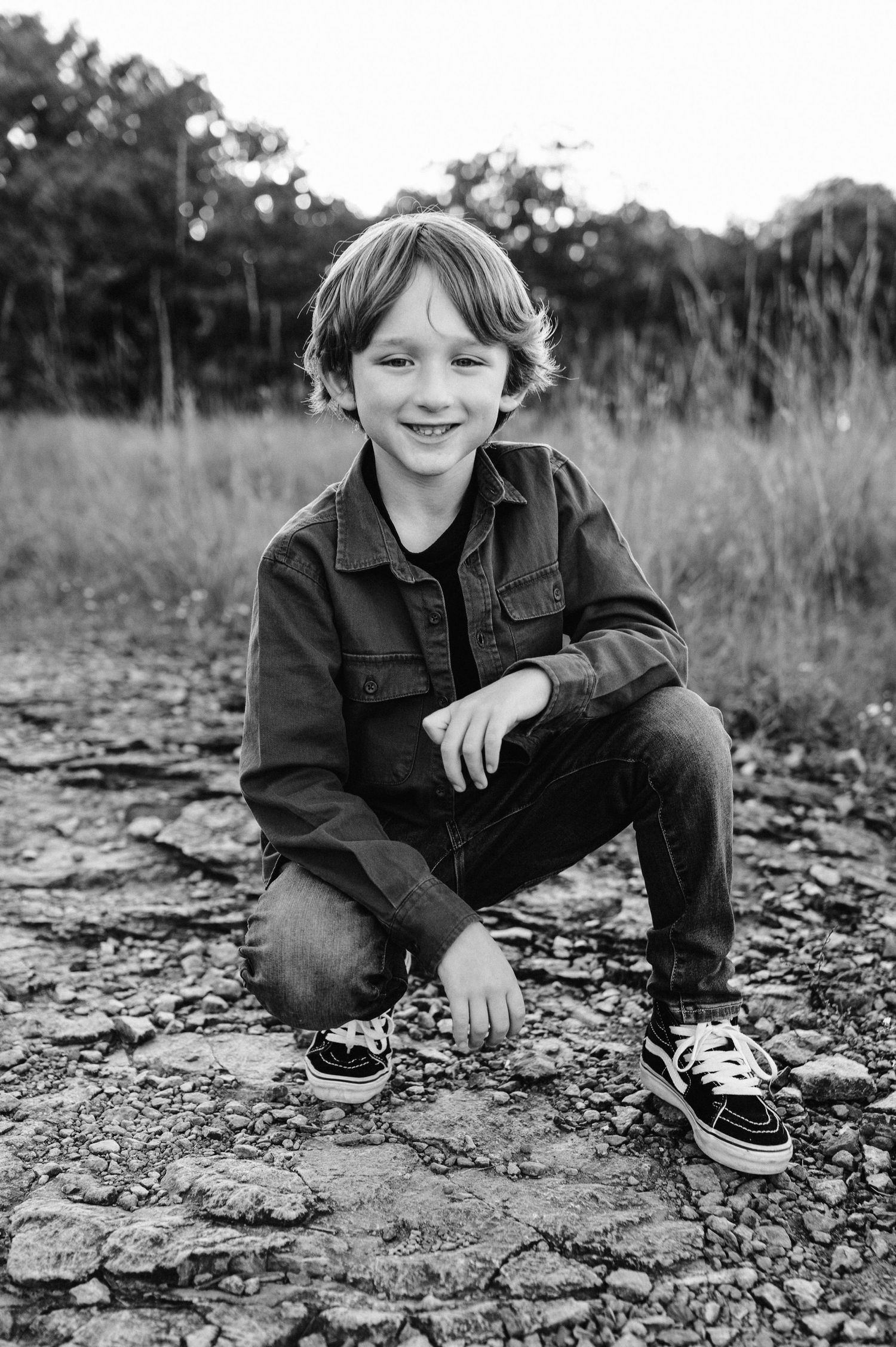 A black and white portrait shows a child crouching on rocky ground wearing casual attire.