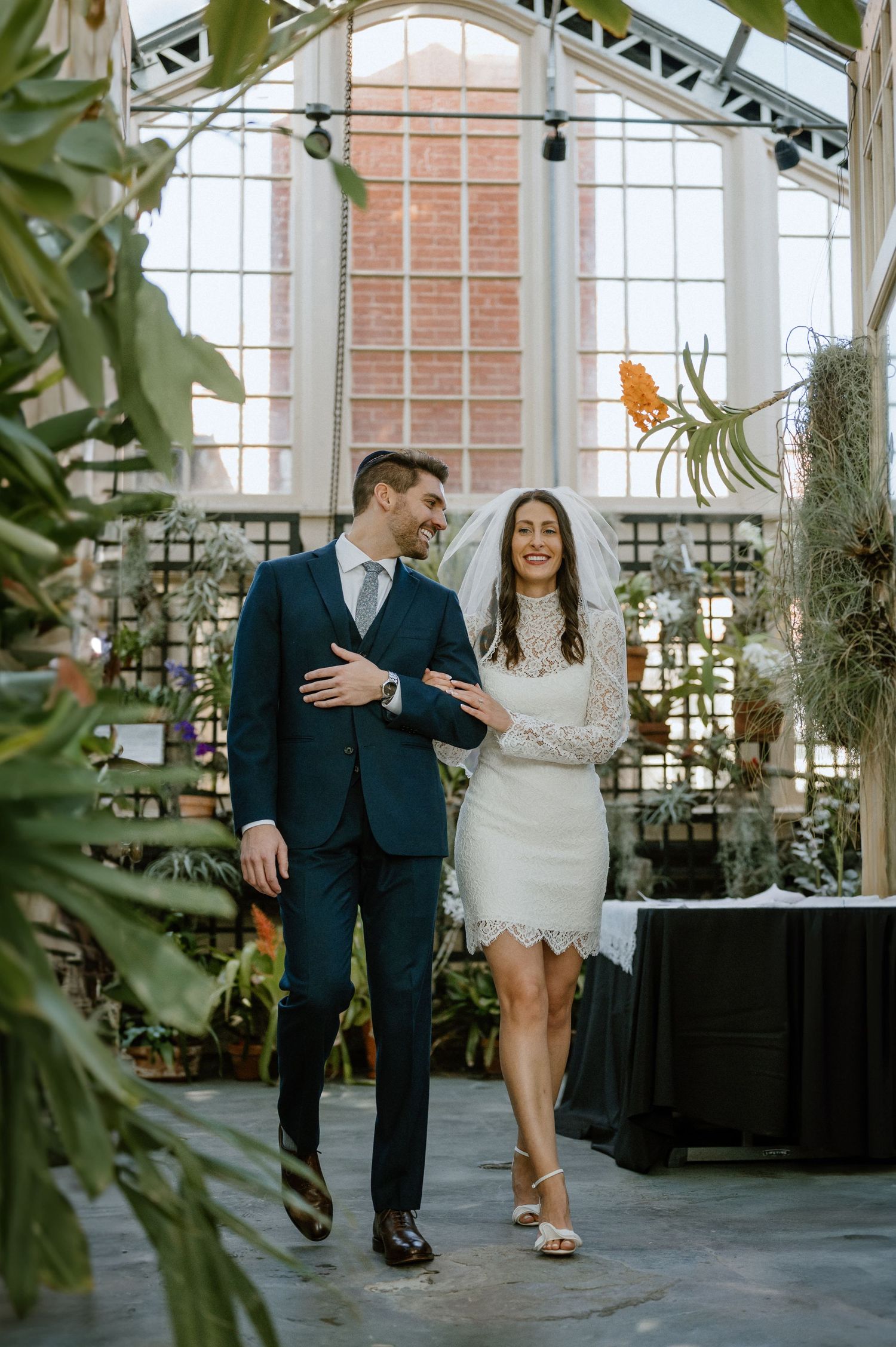 A couple in wedding attire walks through a greenhouse conservatory filled with tropical plants.