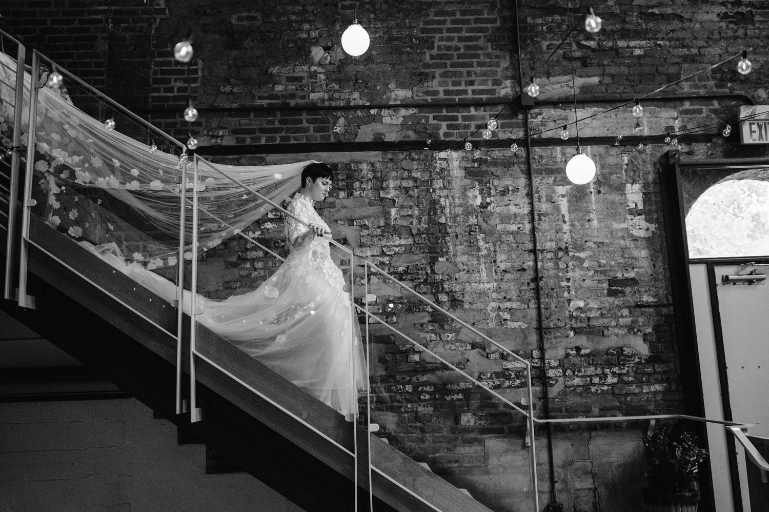 A bride in a flowing white gown descends an industrial staircase with exposed brick walls and string lights.