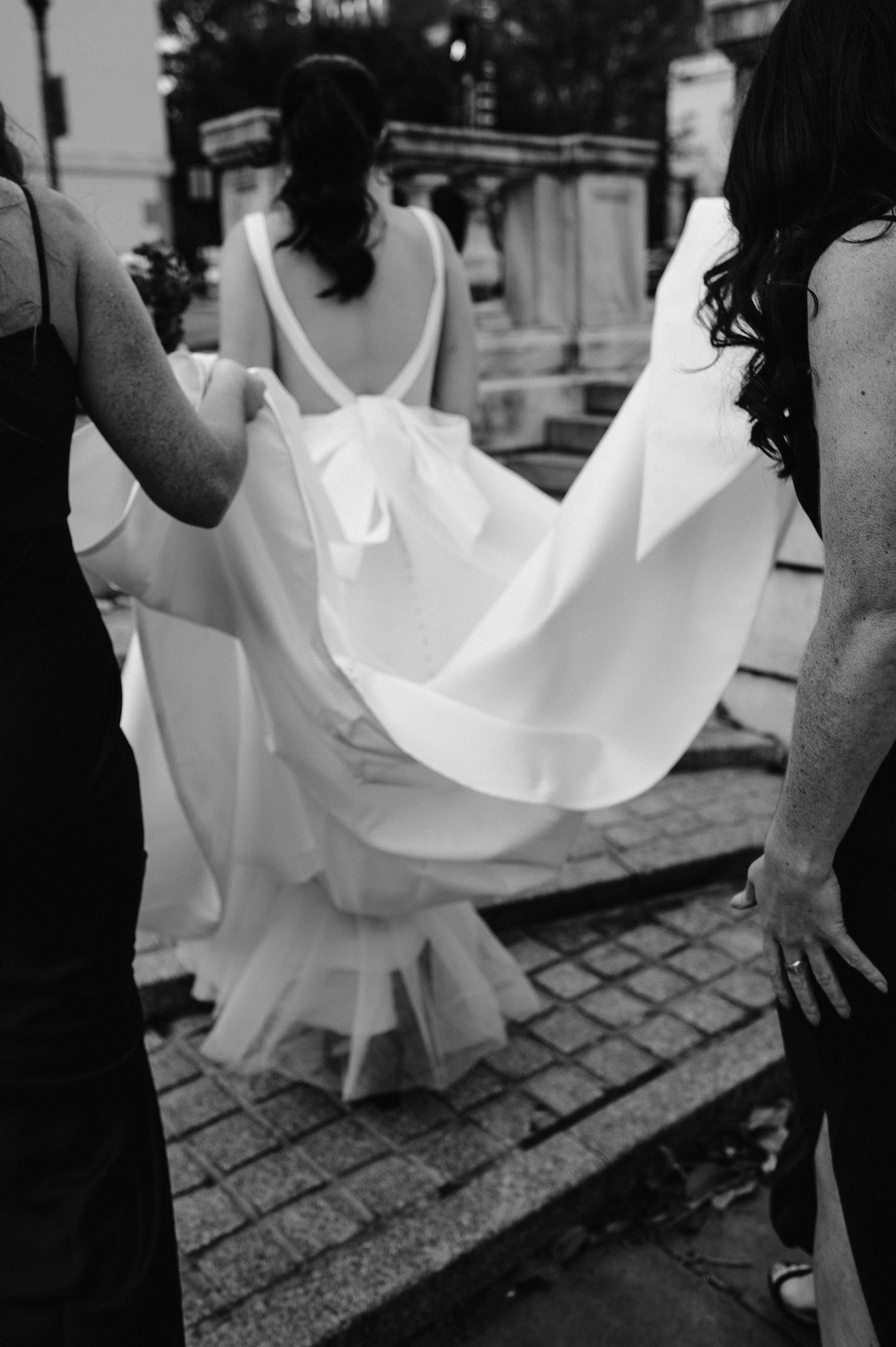 Black and white image of a wedding dress being lifted while walking on cobblestone street.