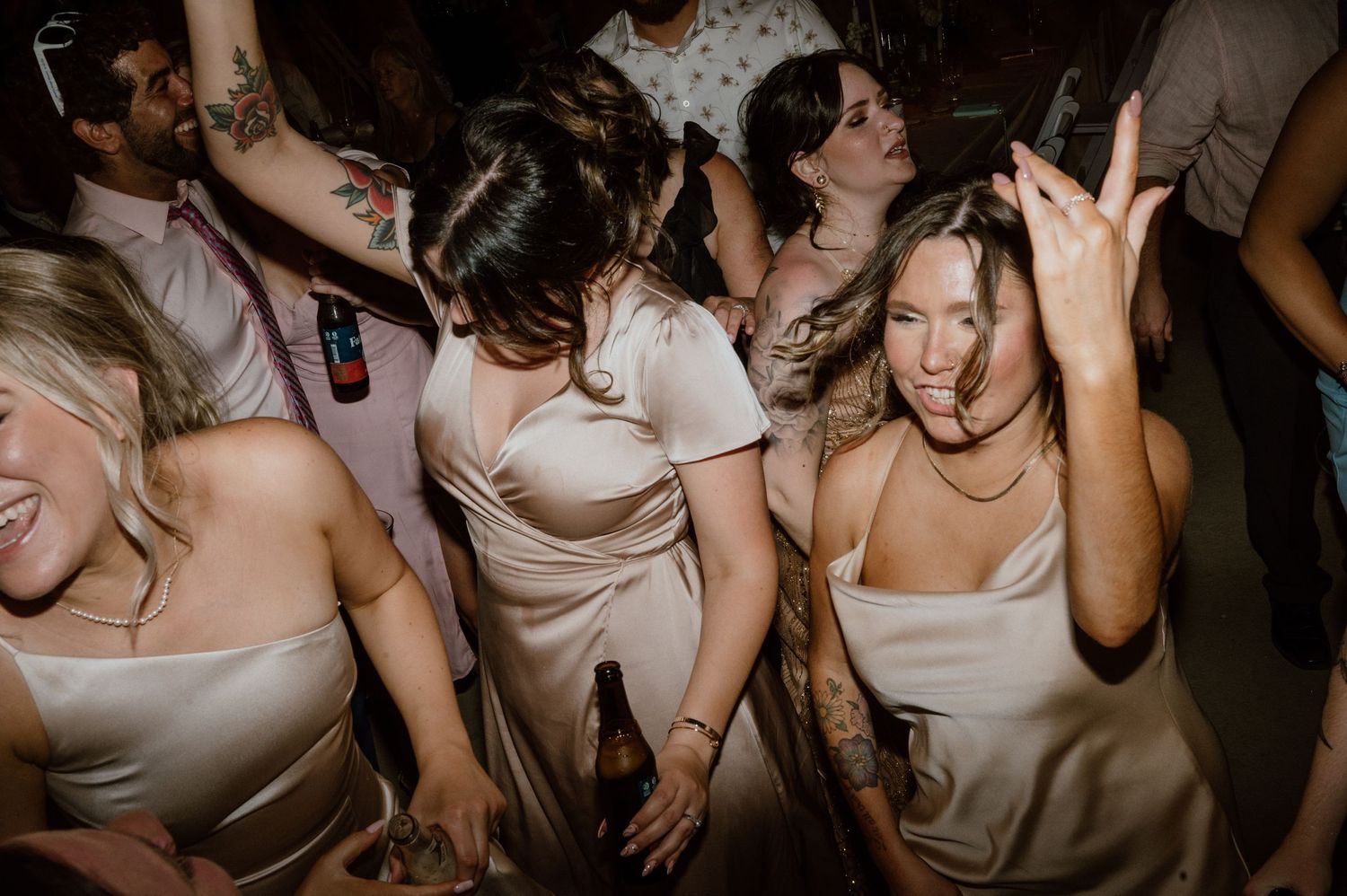 Bridesmaids in beige dresses dancing and celebrating enthusiastically on a dark dance floor.