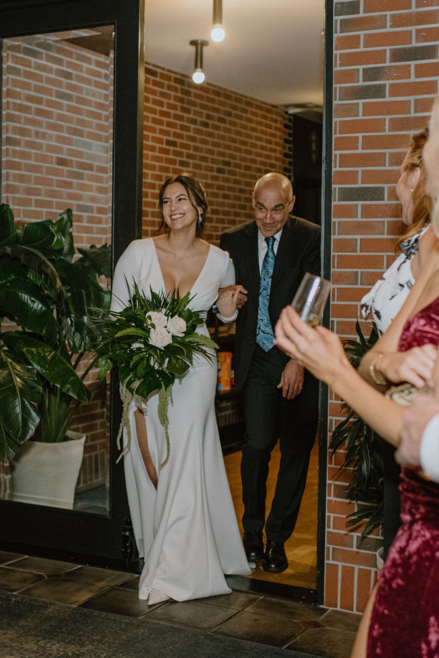 Wedding entrance moment with greenery and brick wall backdrop.