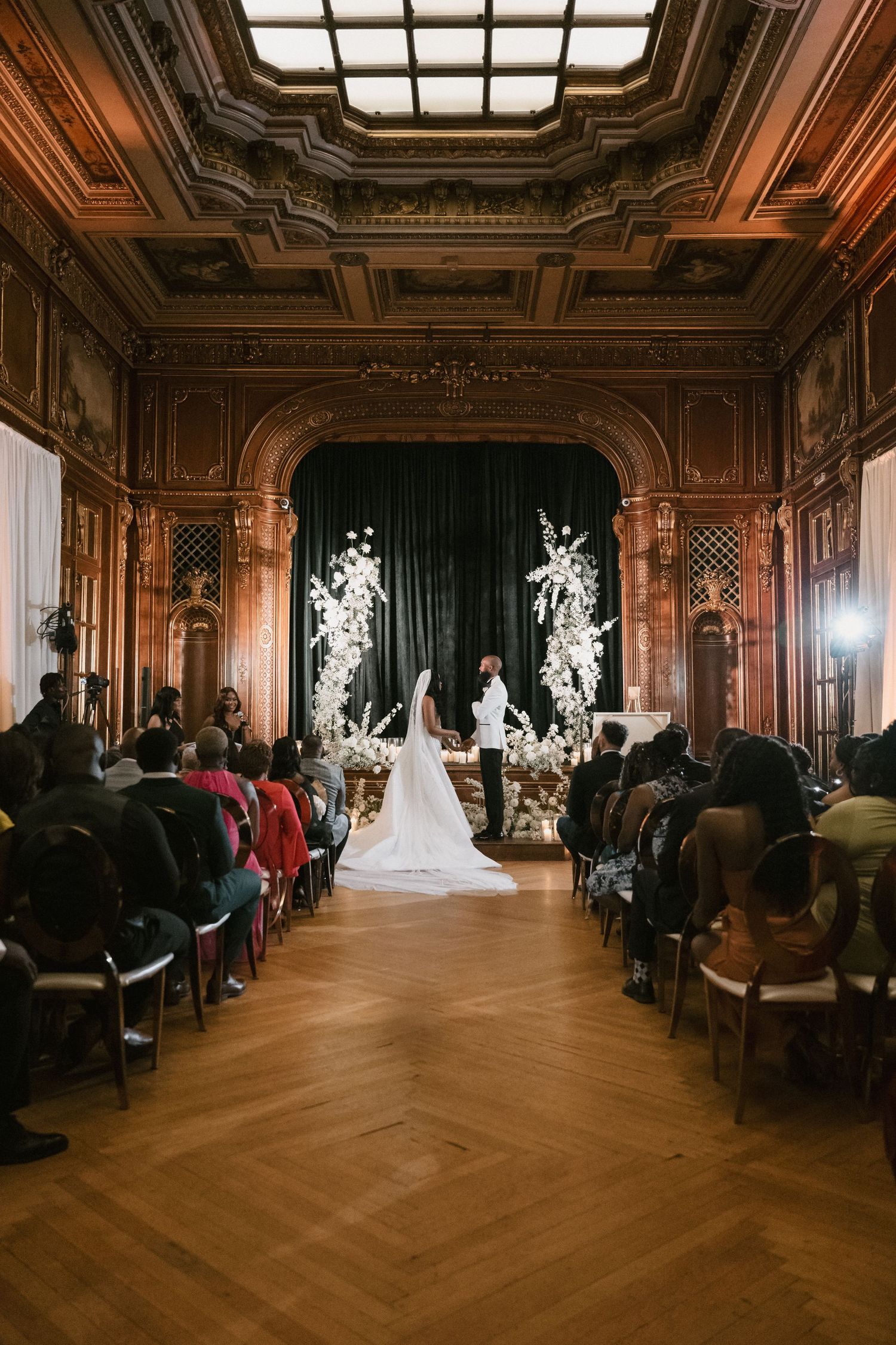 Wedding ceremony in a grand ballroom with ornate wood paneling and guests seated in rows.