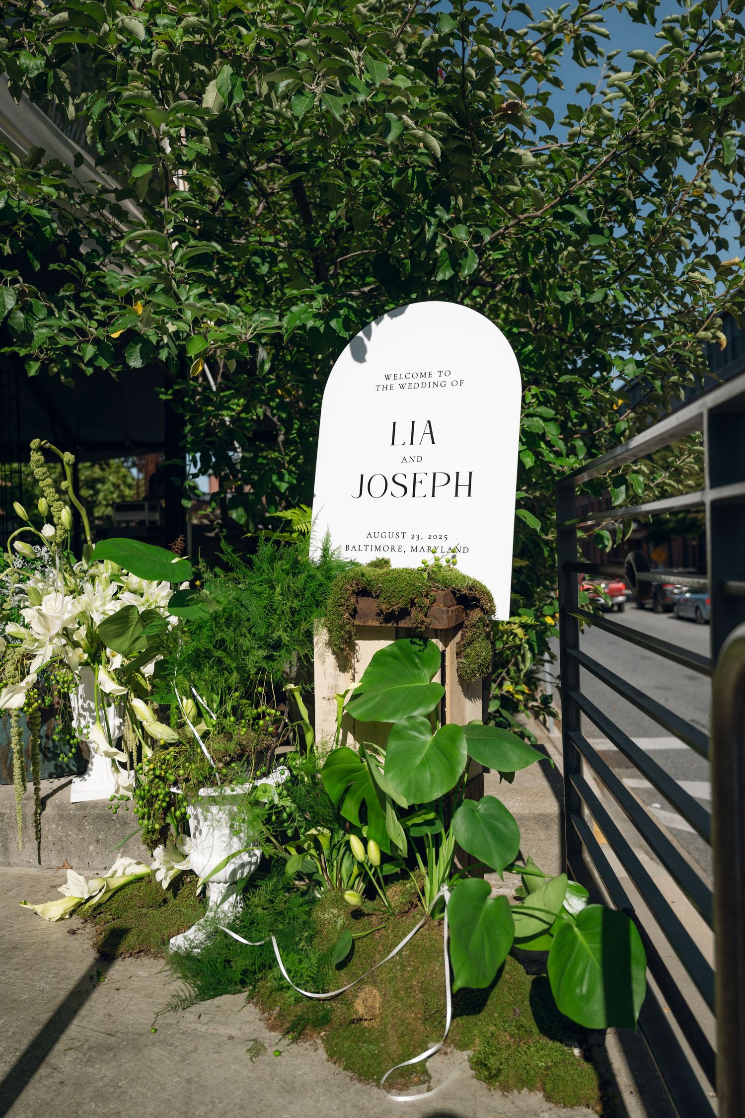 Wedding signage with greenery and white floral decorations against a black railing.