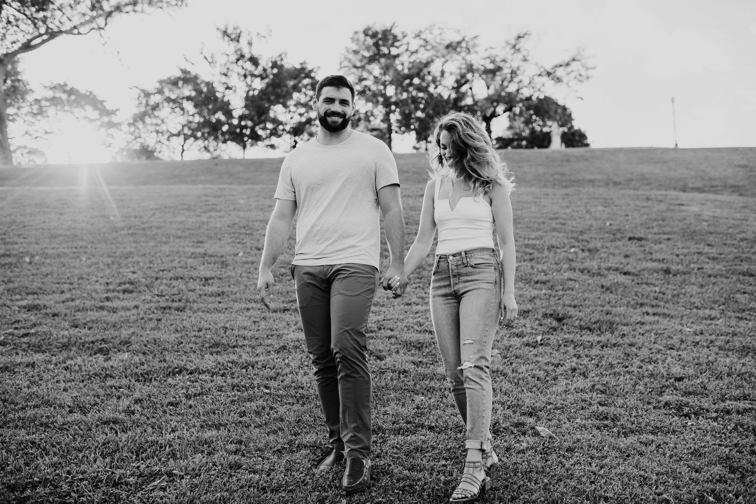Black and white photo of a couple holding hands walking through a sunlit grassy field with trees in the background.
