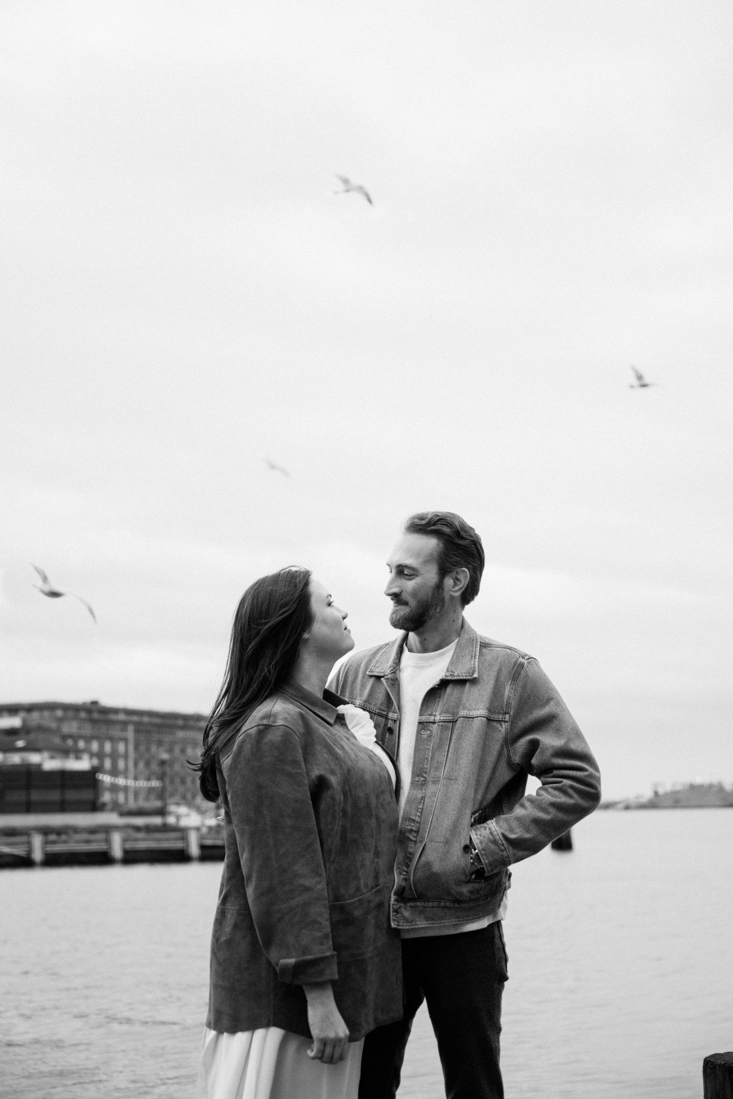 Black and white photo of a romantic couple smiling at each other near a waterfront with seagulls flying overhead.