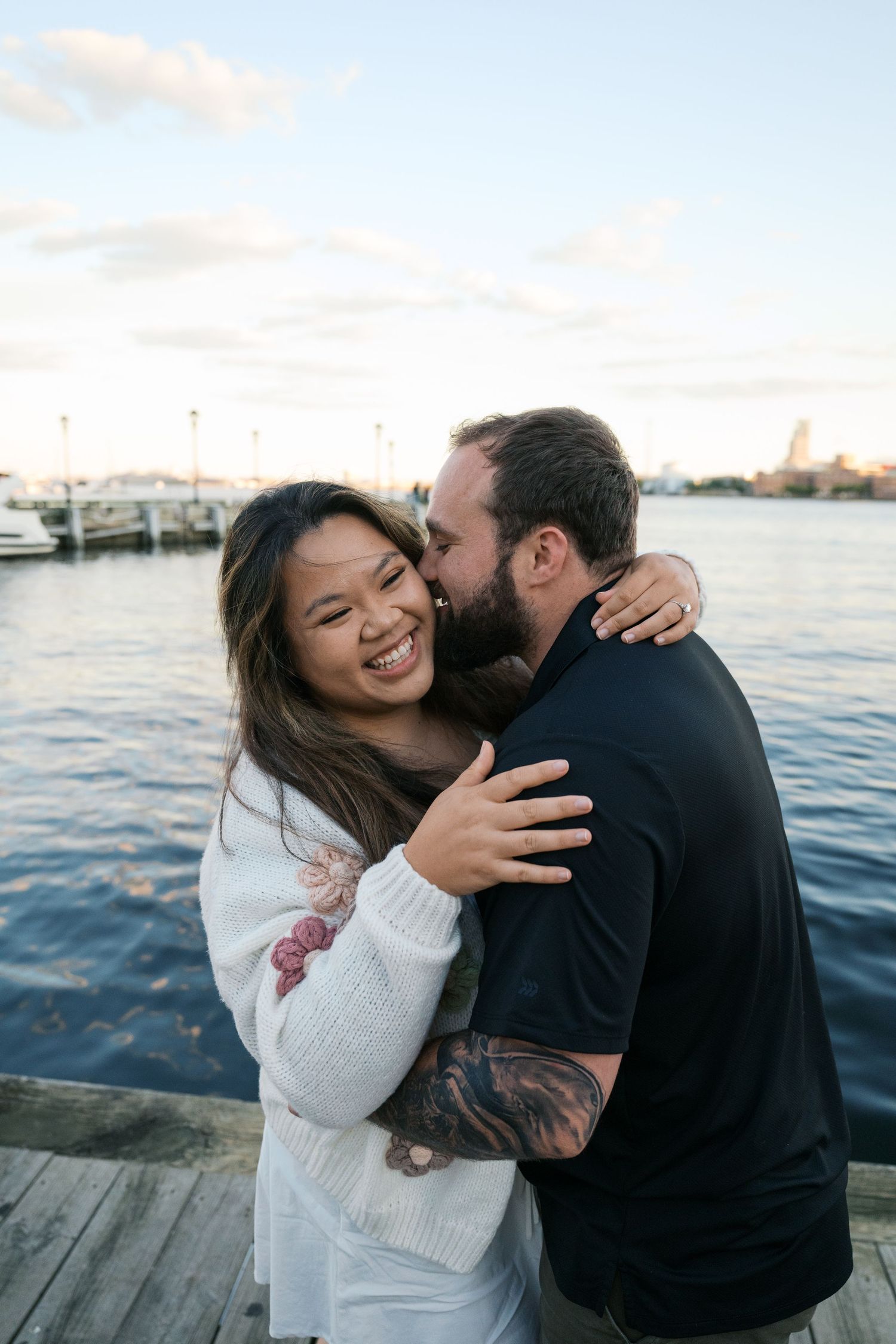 A couple shares a tender kiss on a waterfront dock, woman smiling joyfully, man with tattooed arm embracing her.