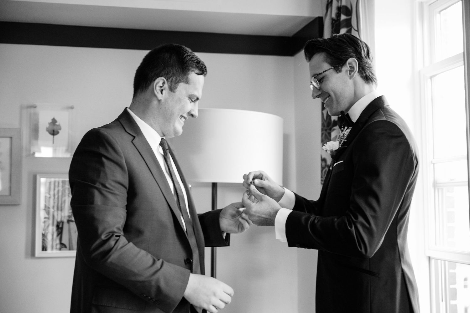 Two men in suits smiling and adjusting cuff links in a bright interior setting.