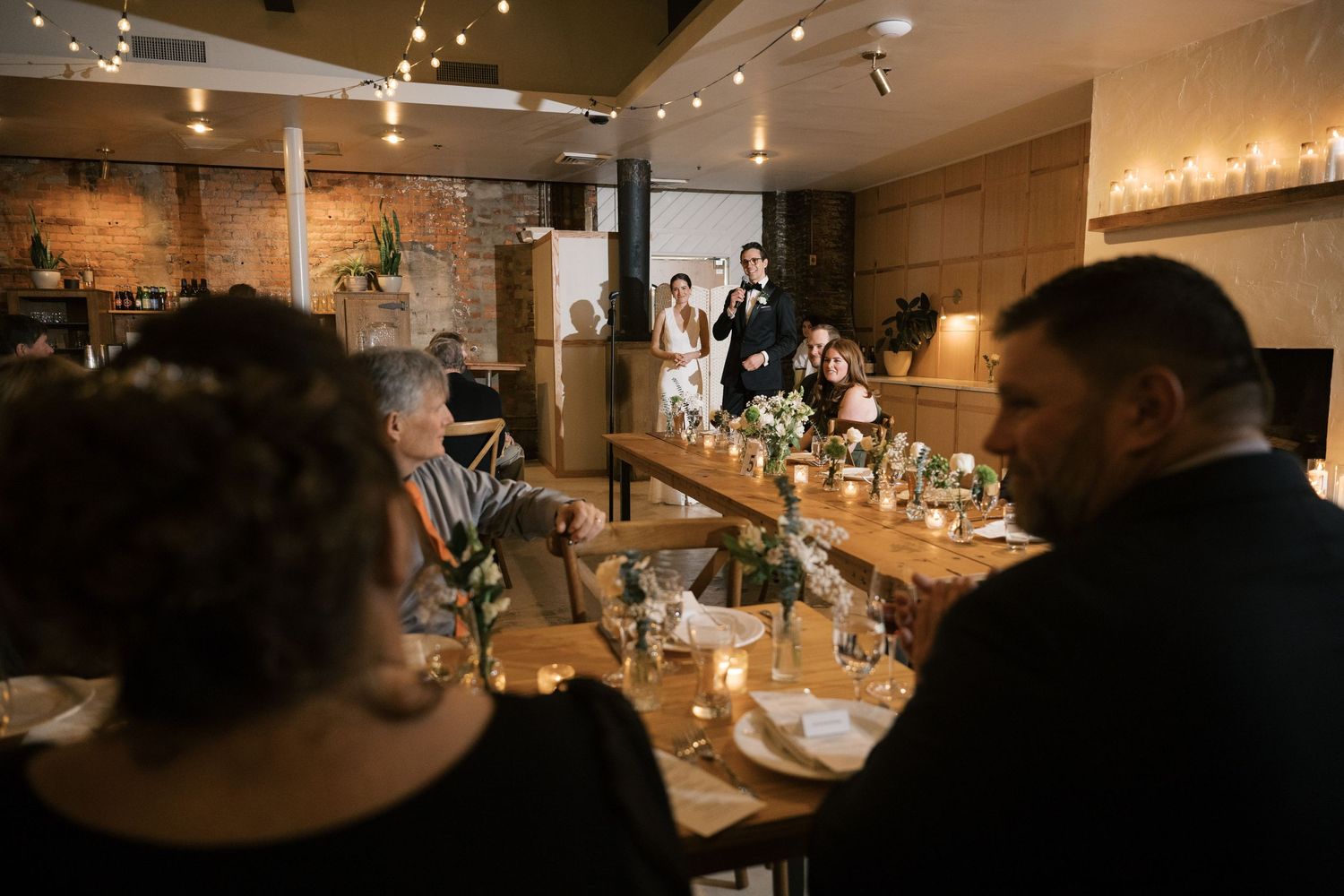 Guests enjoy an intimate dinner setting at a restaurant with ambient lighting and a rustic brick wall interior.