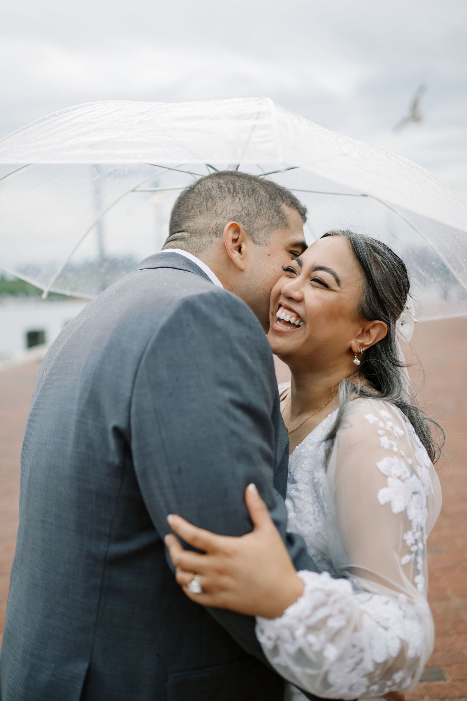 Newlywed couple sharing a joyful moment under a clear umbrella during their rainy wedding day celebration.