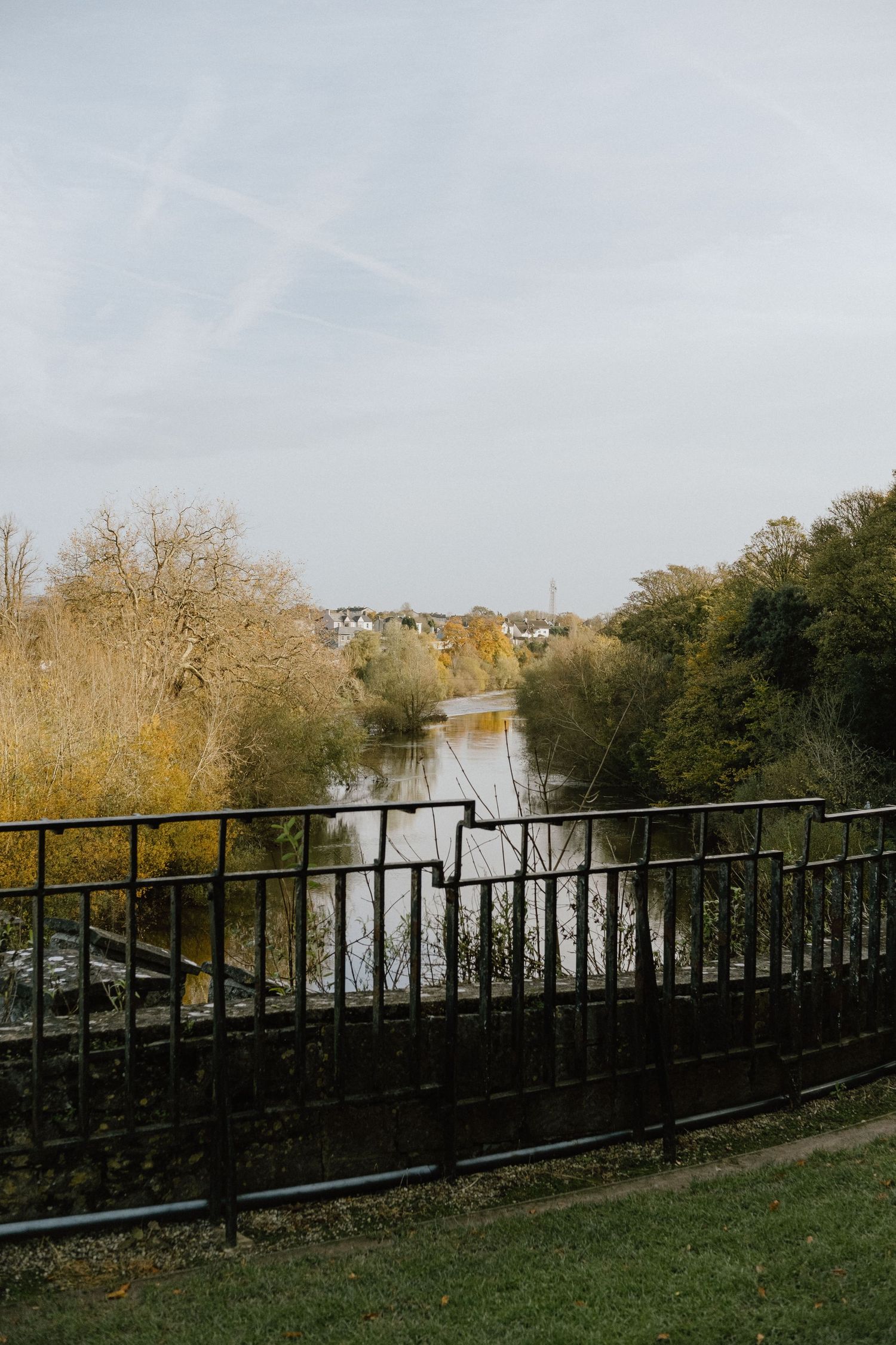 Iron railing overlooks a serene river surrounded by autumn trees on a misty day.