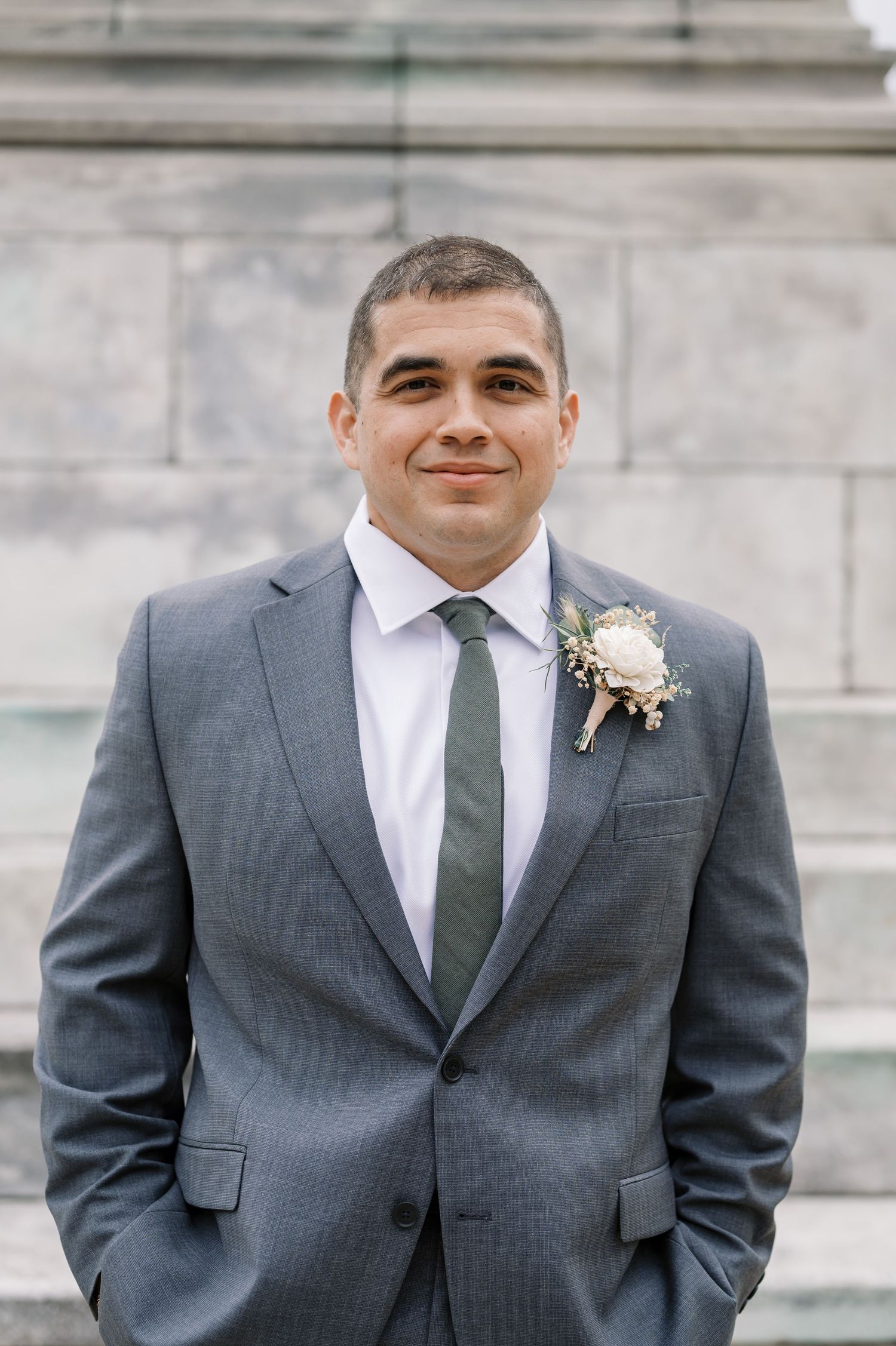 A groom in a grey suit with a boutonniere stands confidently on marble steps in formal wedding attire.