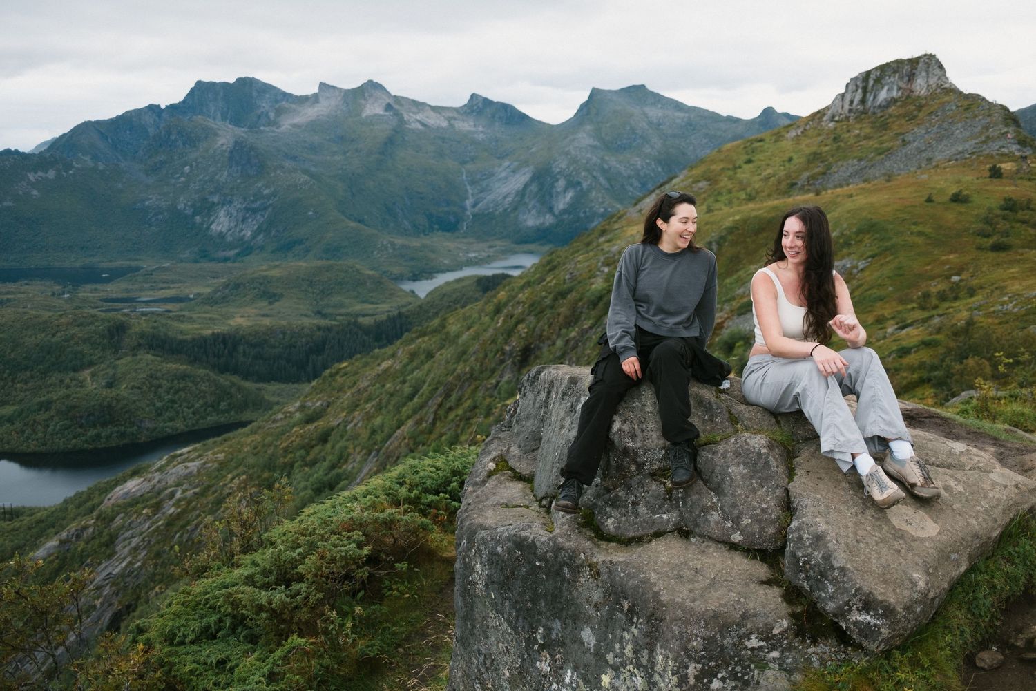 Two figures rest on a large boulder overlooking a mountainous valley with lakes below.