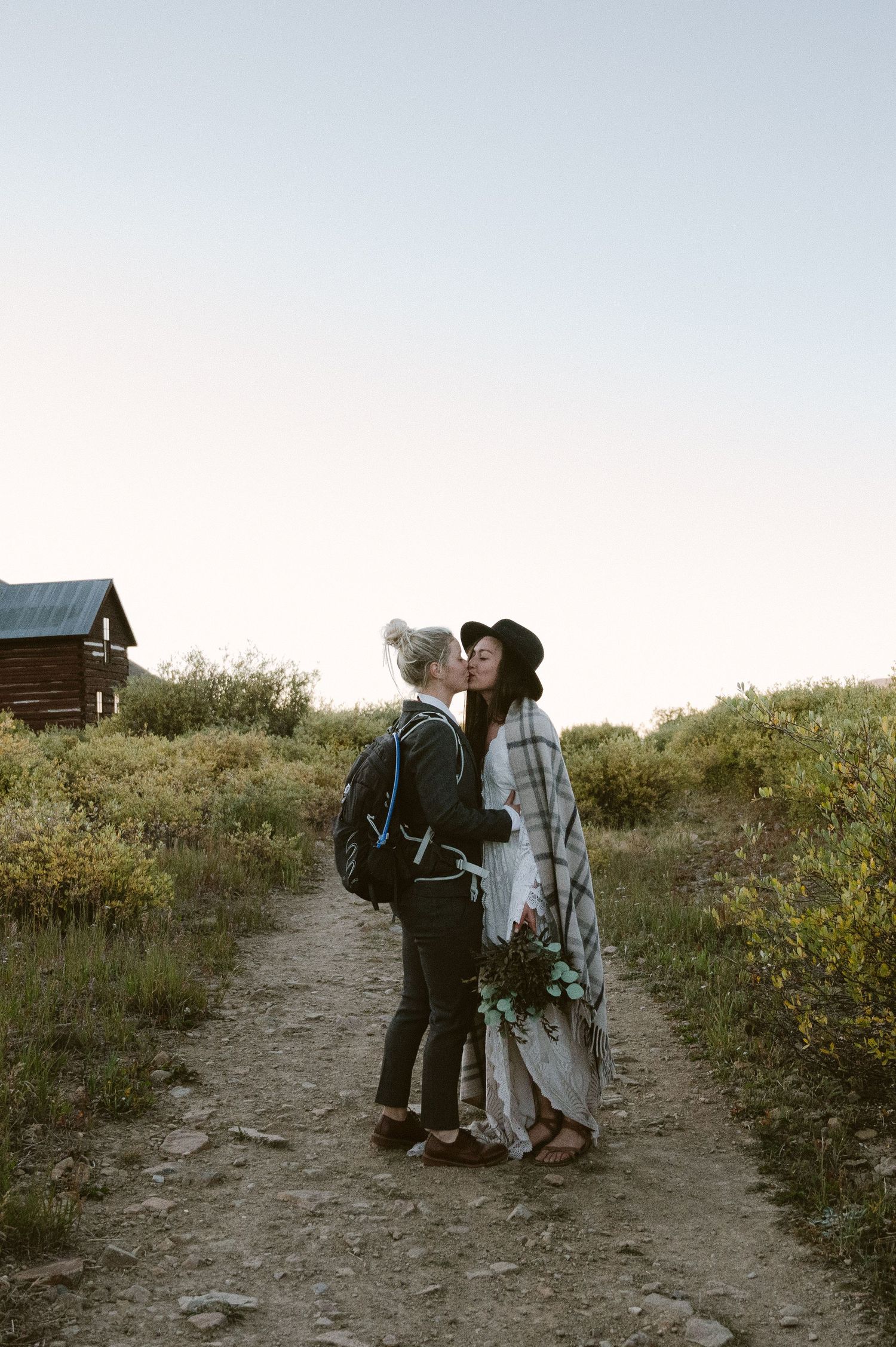 A couple shares a romantic moment on a mountain trail with a rustic cabin in the background.