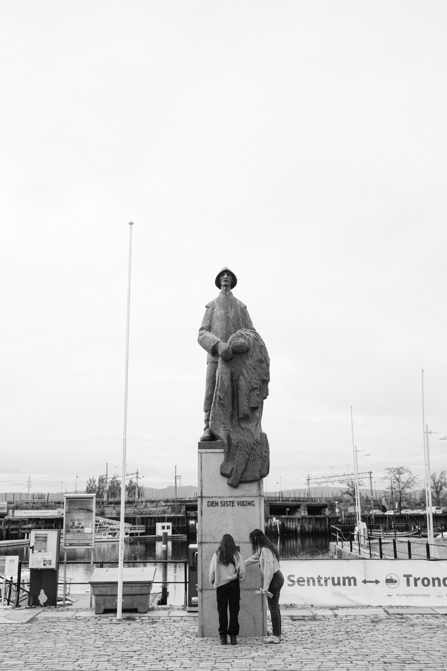 Two people observe a large monument statue in a public square with buildings visible in the background.