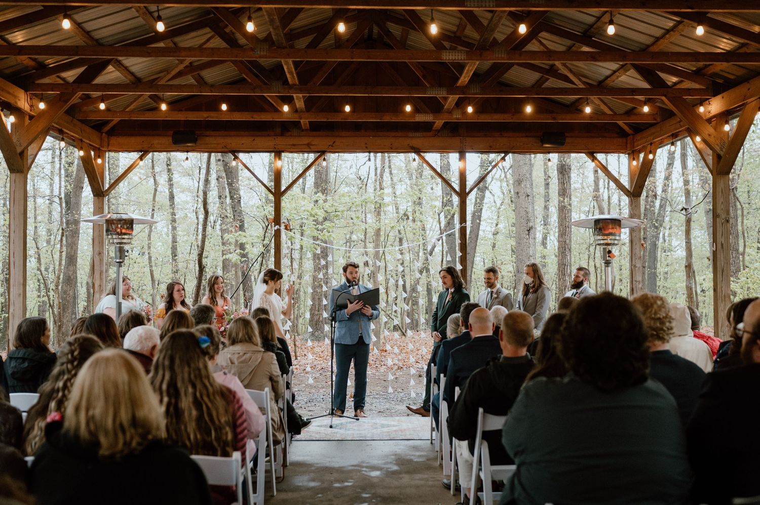 Rustic woodland wedding ceremony under covered pavilion surrounded by trees.