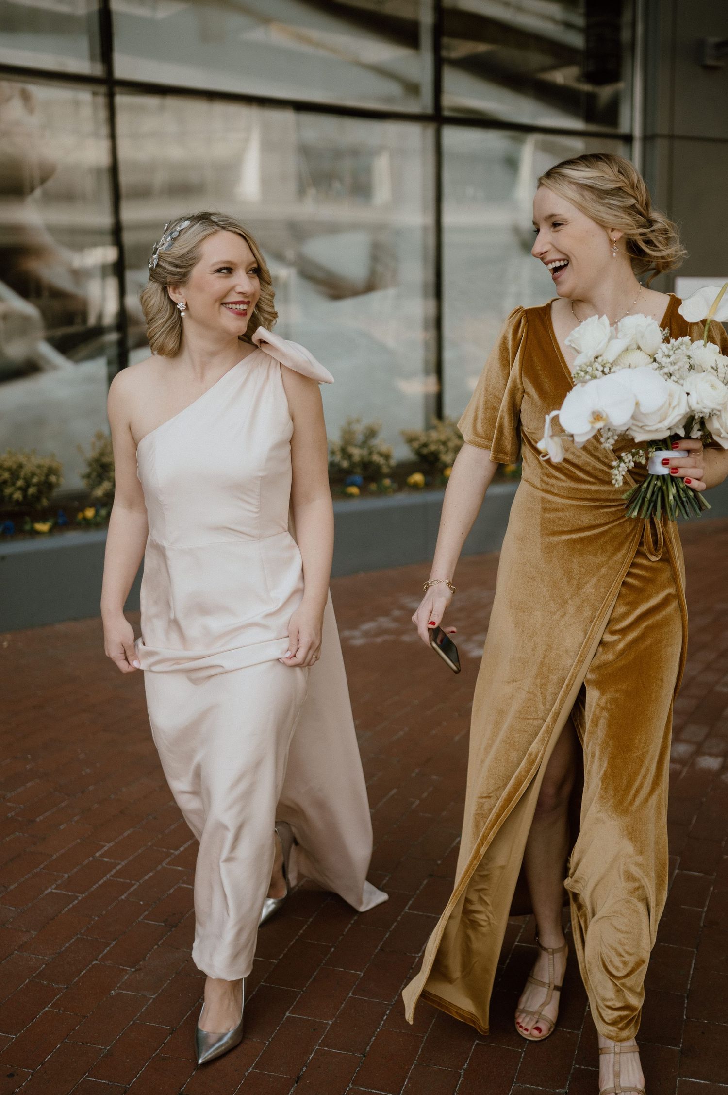 Two bridesmaids in blush and golden velvet dresses walk together while smiling.