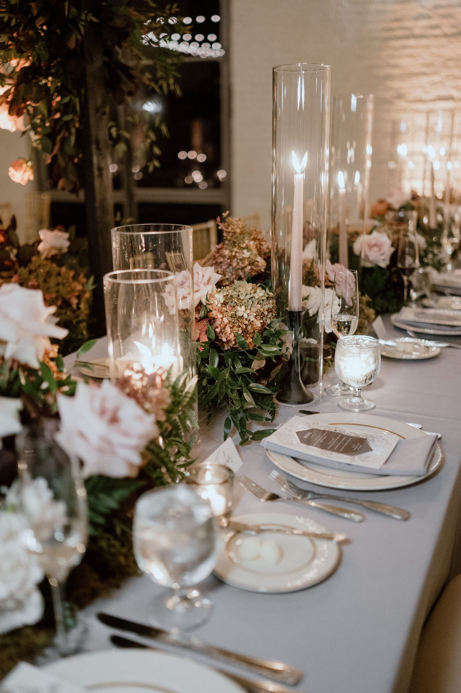 Romantic wedding table setting with tall glass candleholders, lush greenery and soft floral arrangements.