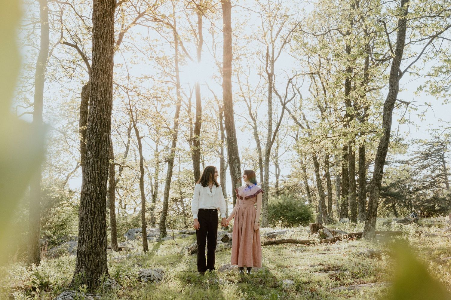 Two figures stand together in a sunlit forest with golden rays filtering through the trees.