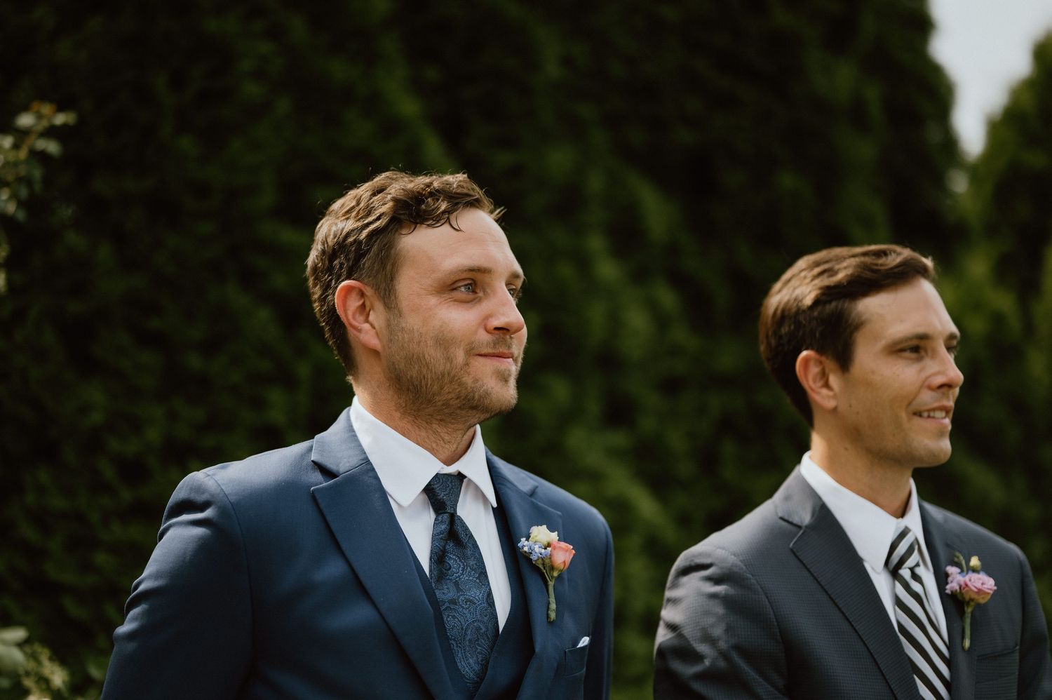 Two groomsmen in navy suits stand together during an outdoor wedding.