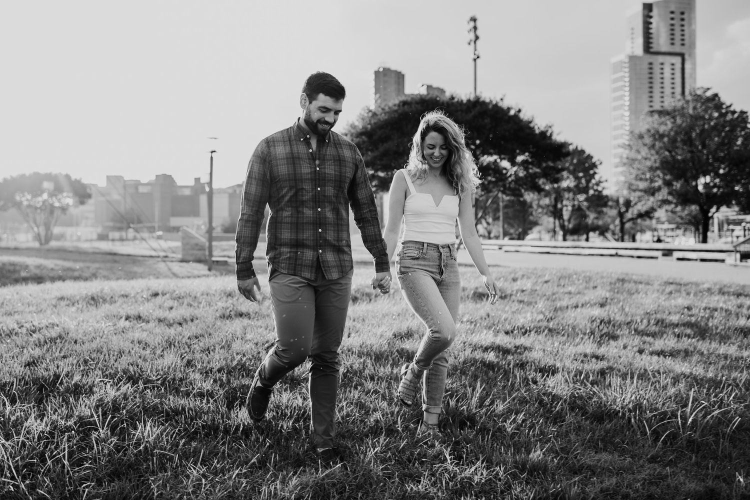 Black and white photo of couple walking through grassy field with city backdrop.