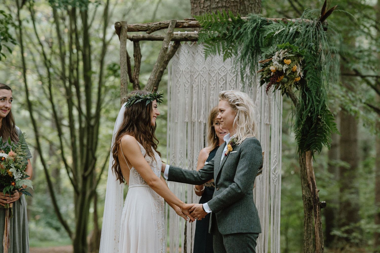 Couple holds hands during their forest wedding ceremony under a decorative arch.