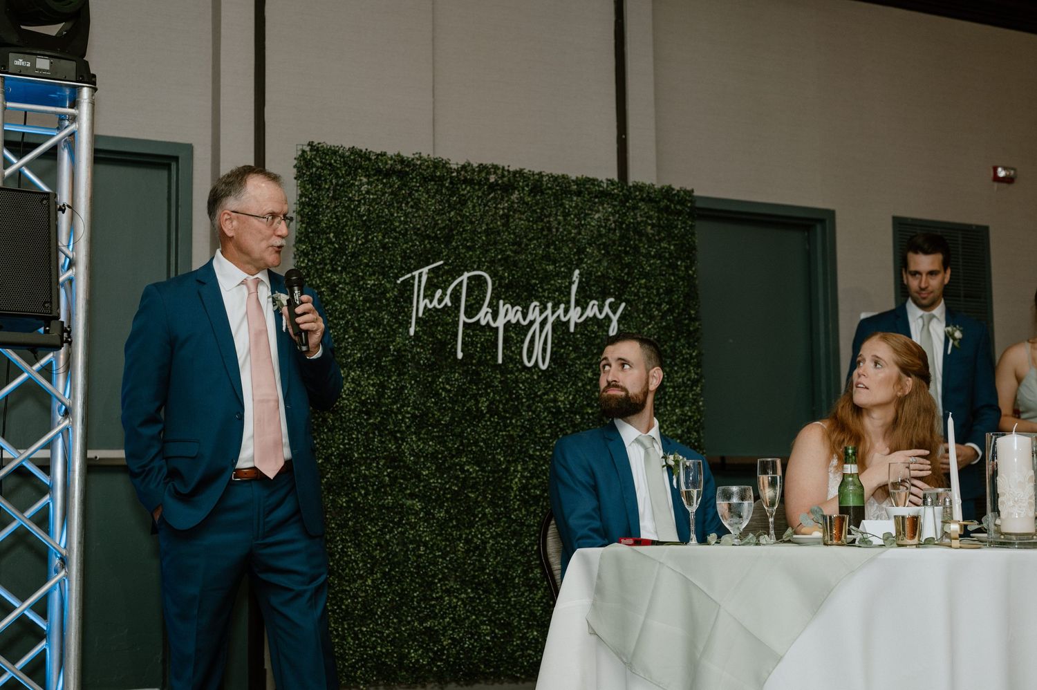 Wedding reception scene with speakers giving toasts in front of a hedge wall backdrop.