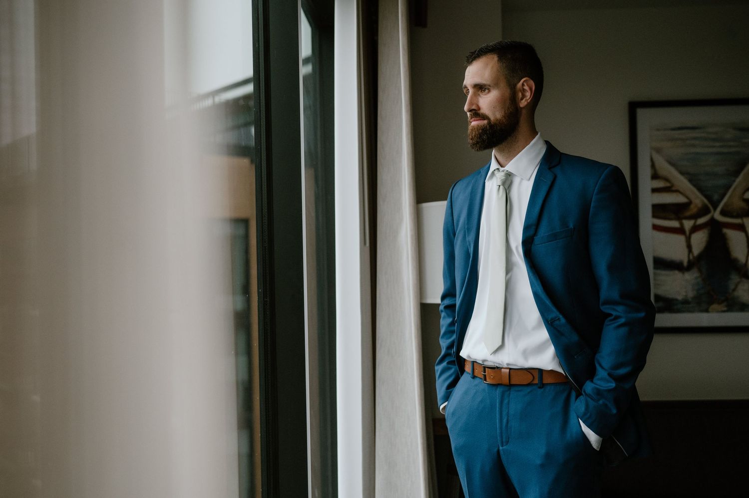 Person wearing blue suit standing by window in modern room with hands in pockets.