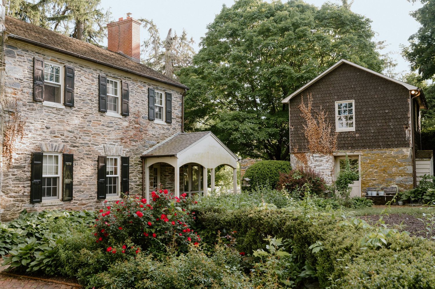 Historic stone farmhouse with black shutters and a connecting walkway to smaller outbuilding.