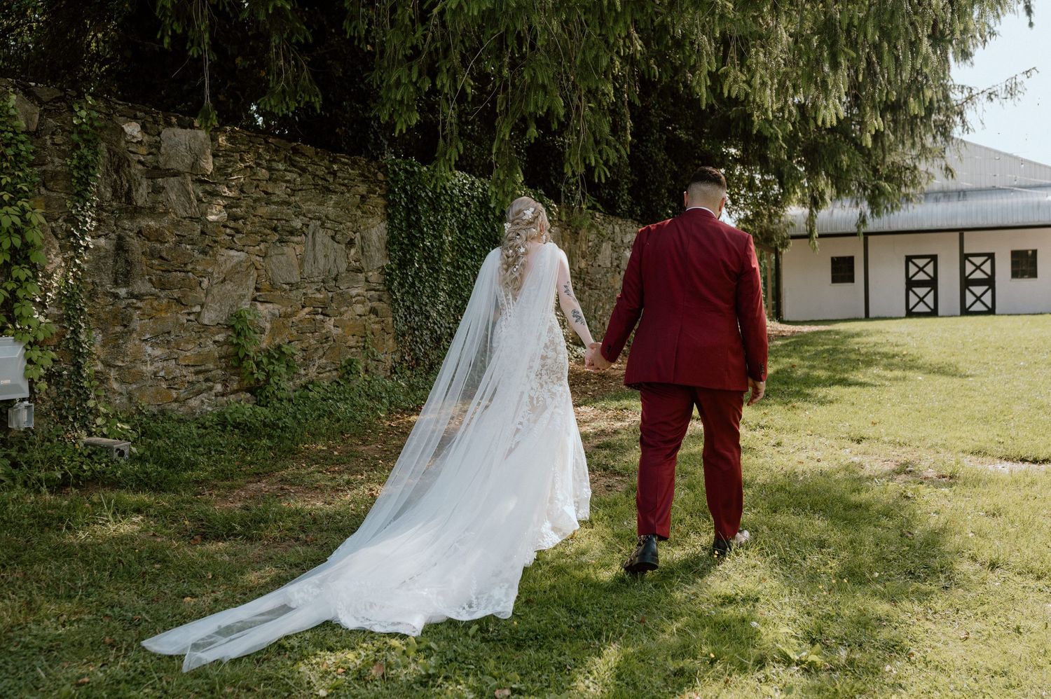 A couple walks on grass near a stone wall, with the bride wearing a flowing white gown and long veil.