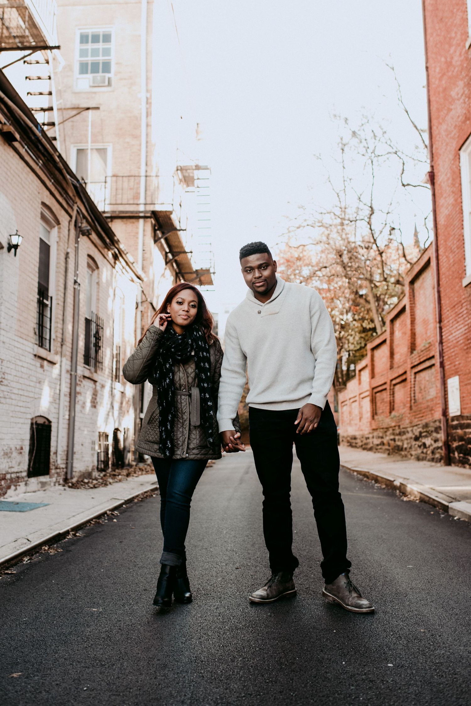 A stylish couple posing in an urban setting with brick buildings and autumn trees.