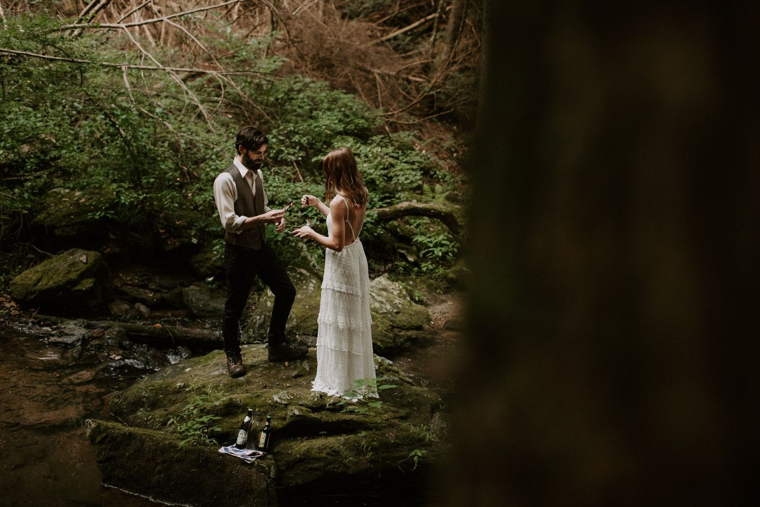 A couple shares a moment while standing on mossy rocks in a wooded setting.