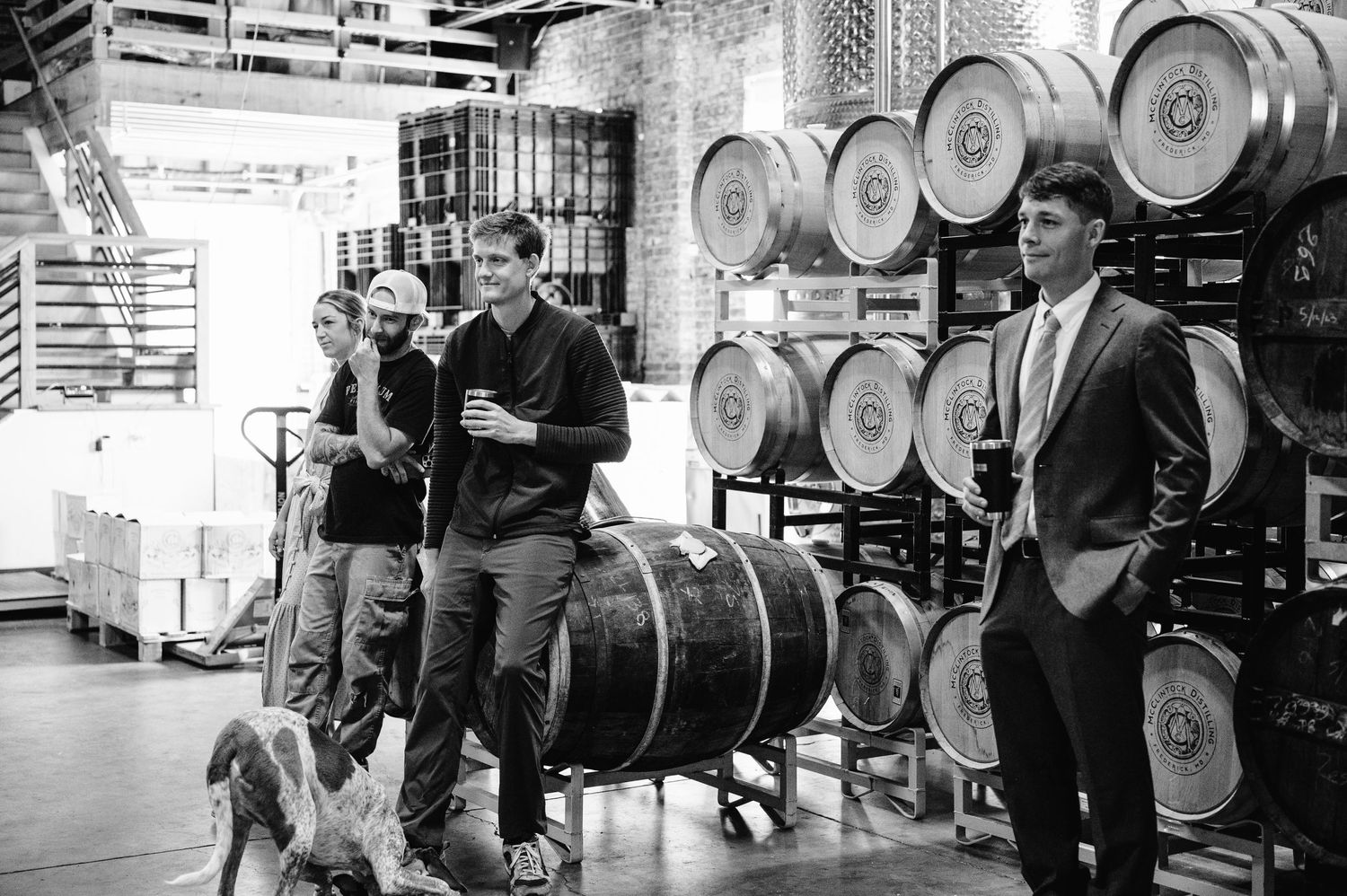 Black and white photo of people gathered among whiskey barrels in a distillery.