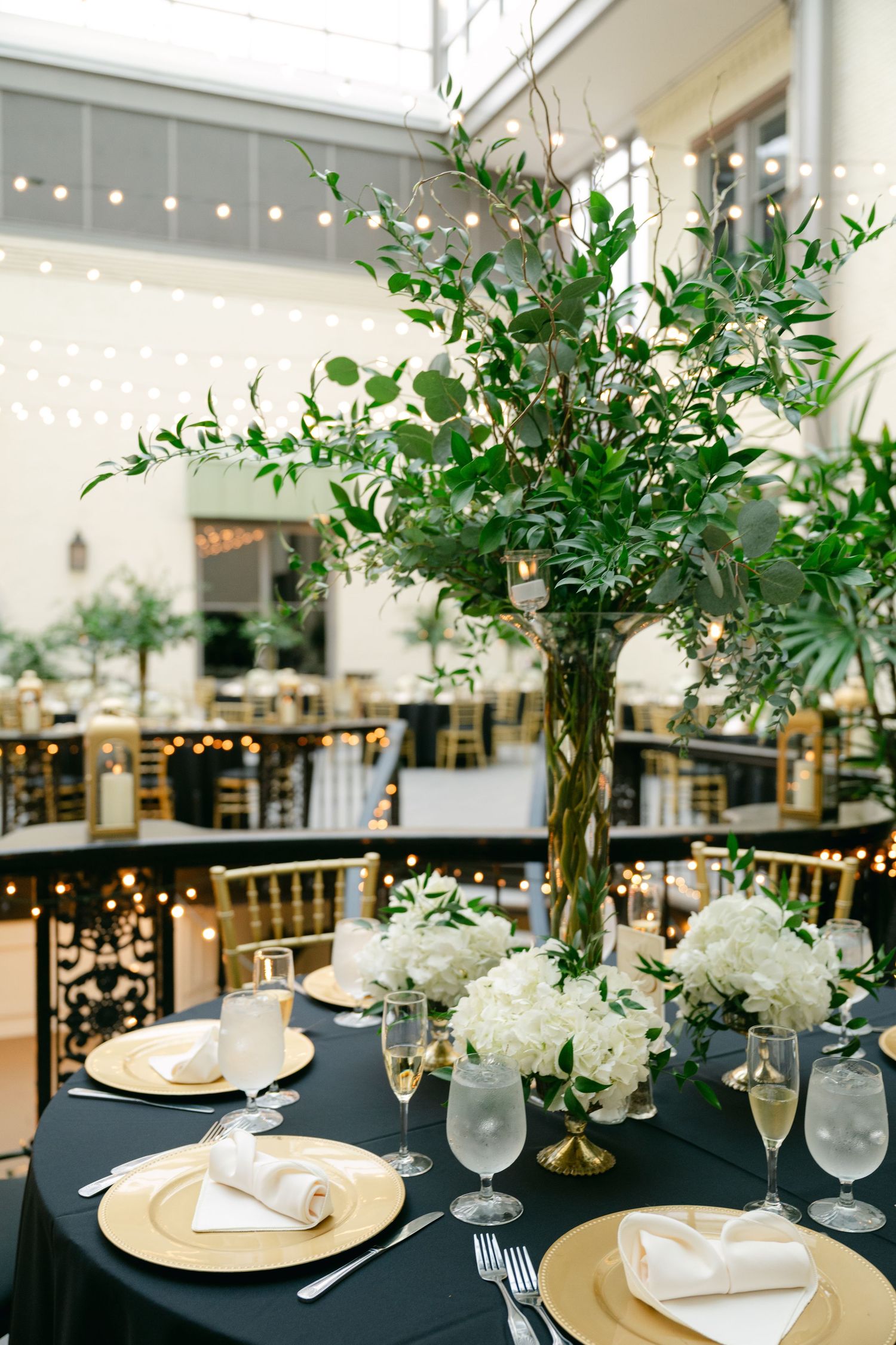 Elegant wedding reception table with tall greenery centerpiece, gold chargers and string lights overhead.