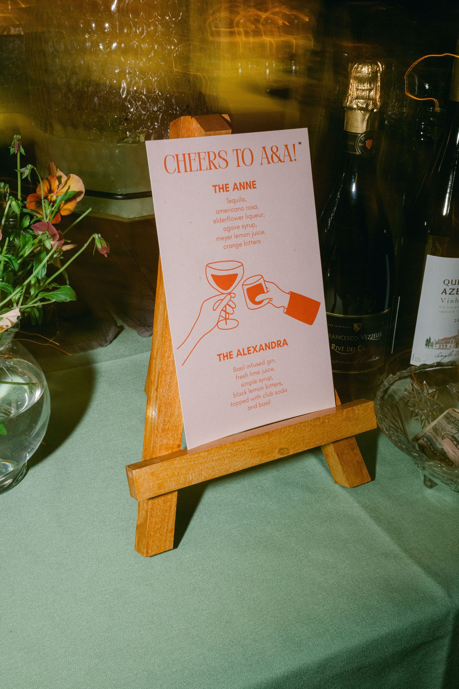 Sign displays drink menu choices on wooden stands with 'Cheers' text at an event table.