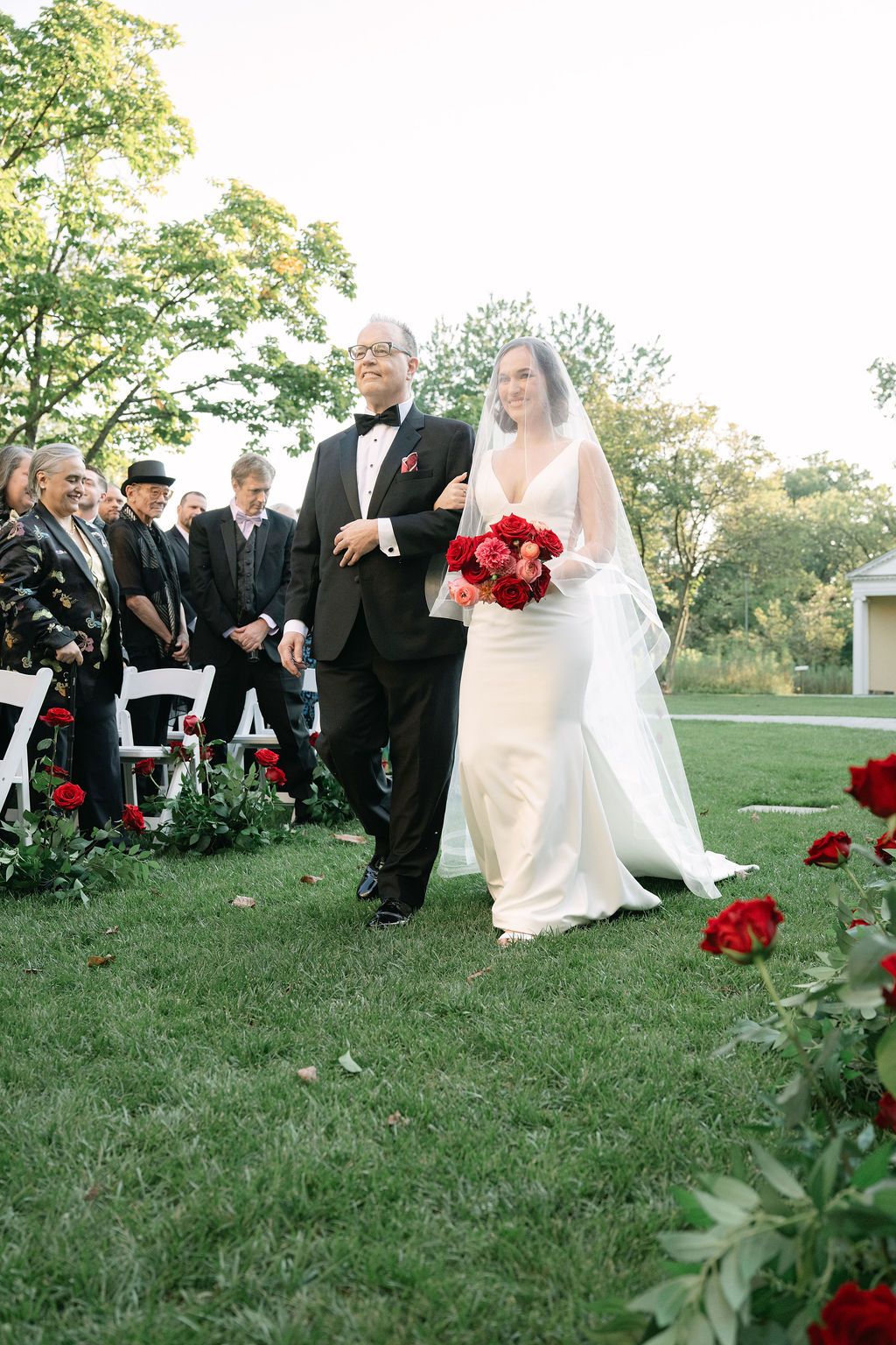 A wedding ceremony takes place outdoors on a grassy lawn with red roses and guests seated in formal attire.