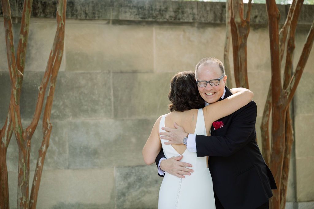 A wedding couple embraces in front of a textured concrete wall with crepe myrtle trees creating a romantic outdoor setting.