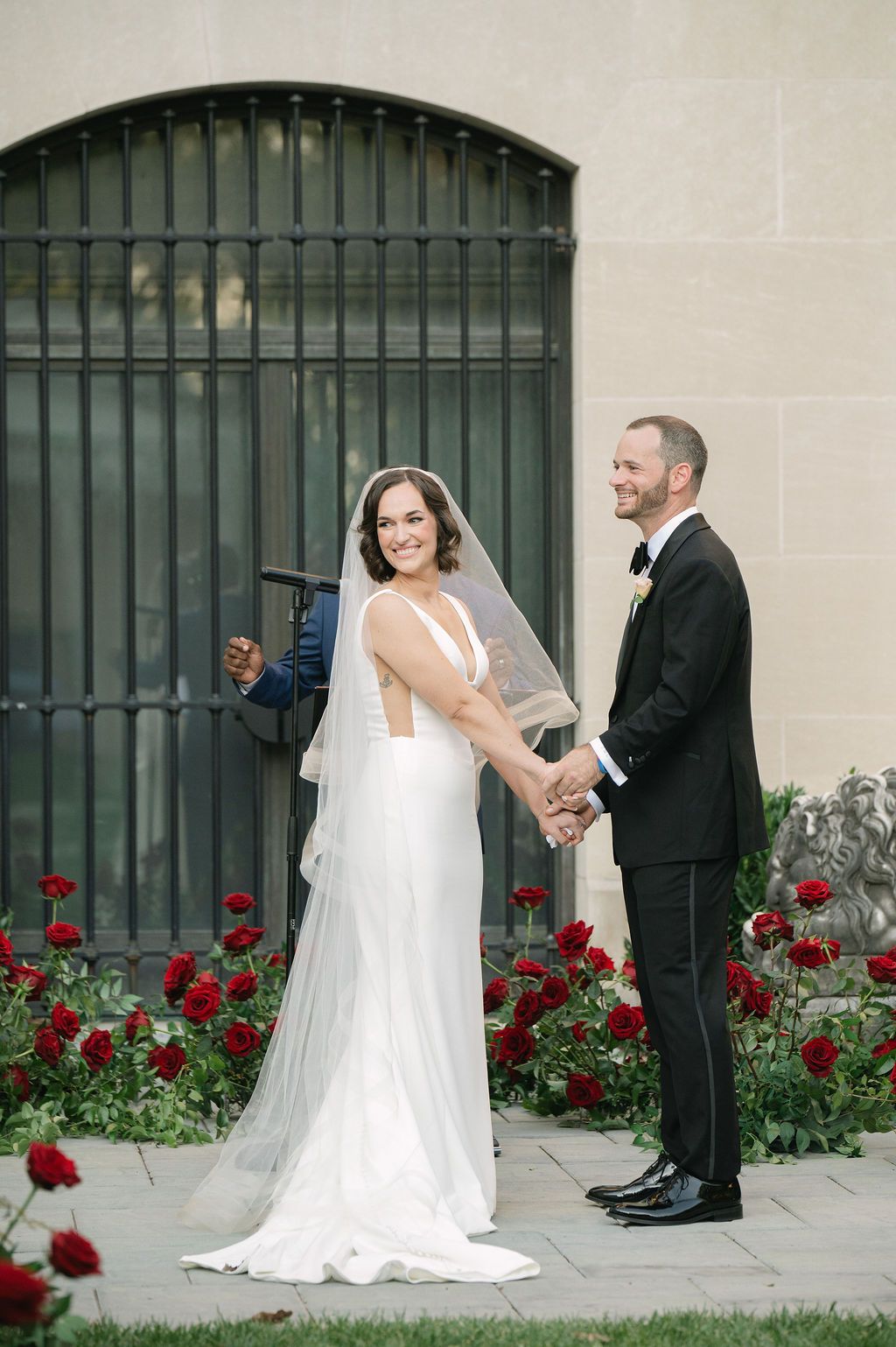 A wedding couple poses in front of a green metal gate with red roses blooming along a stone wall.