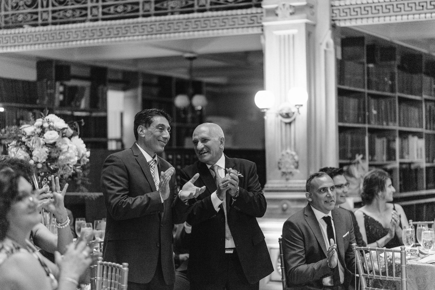 Wedding guests raise glasses during speeches in an elegant library venue with ornate columns and bookshelves.