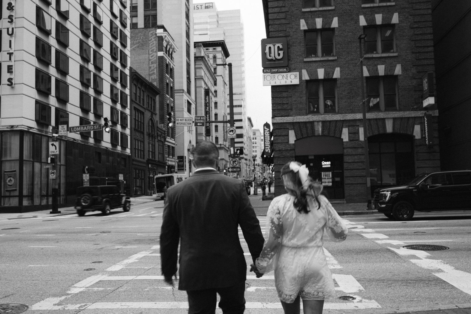 A couple in wedding attire walks hand in hand down a city street in black and white.