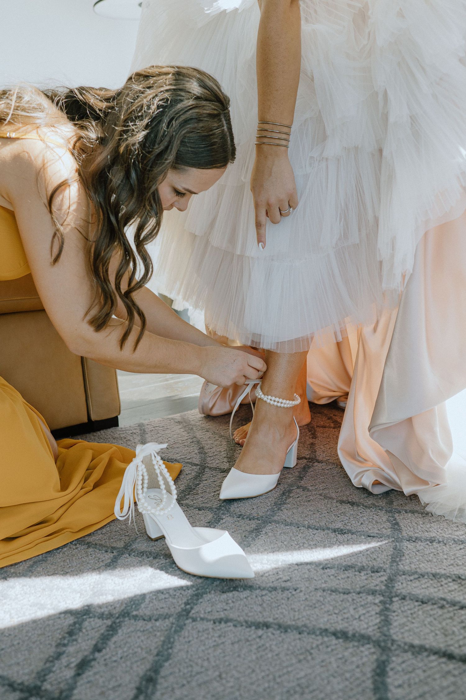 Helper fastening bride's shoe during wedding preparations in sunlit room.