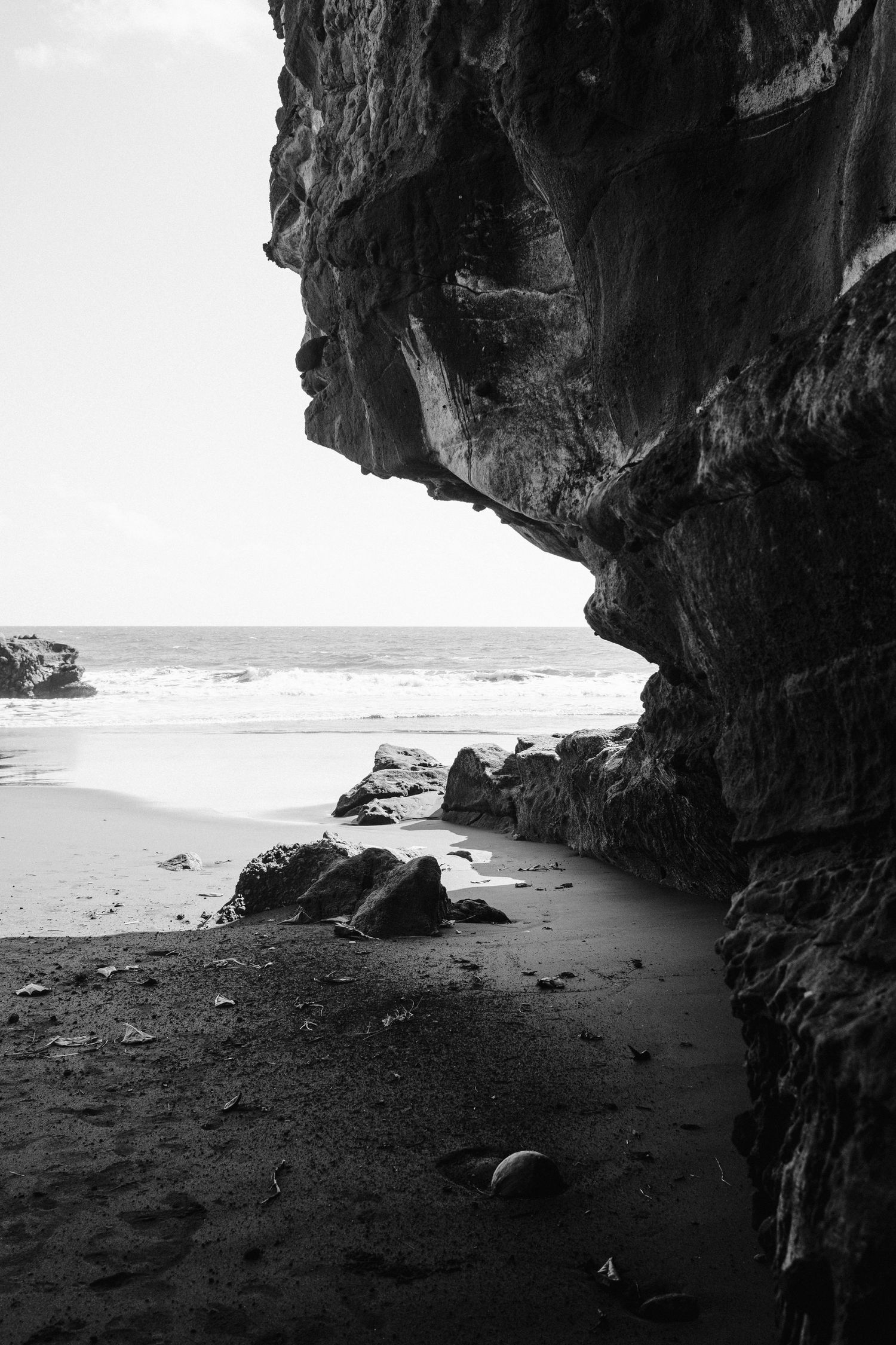 Dramatic black and white view through a seaside cave entrance showing rocky beach and crashing waves.