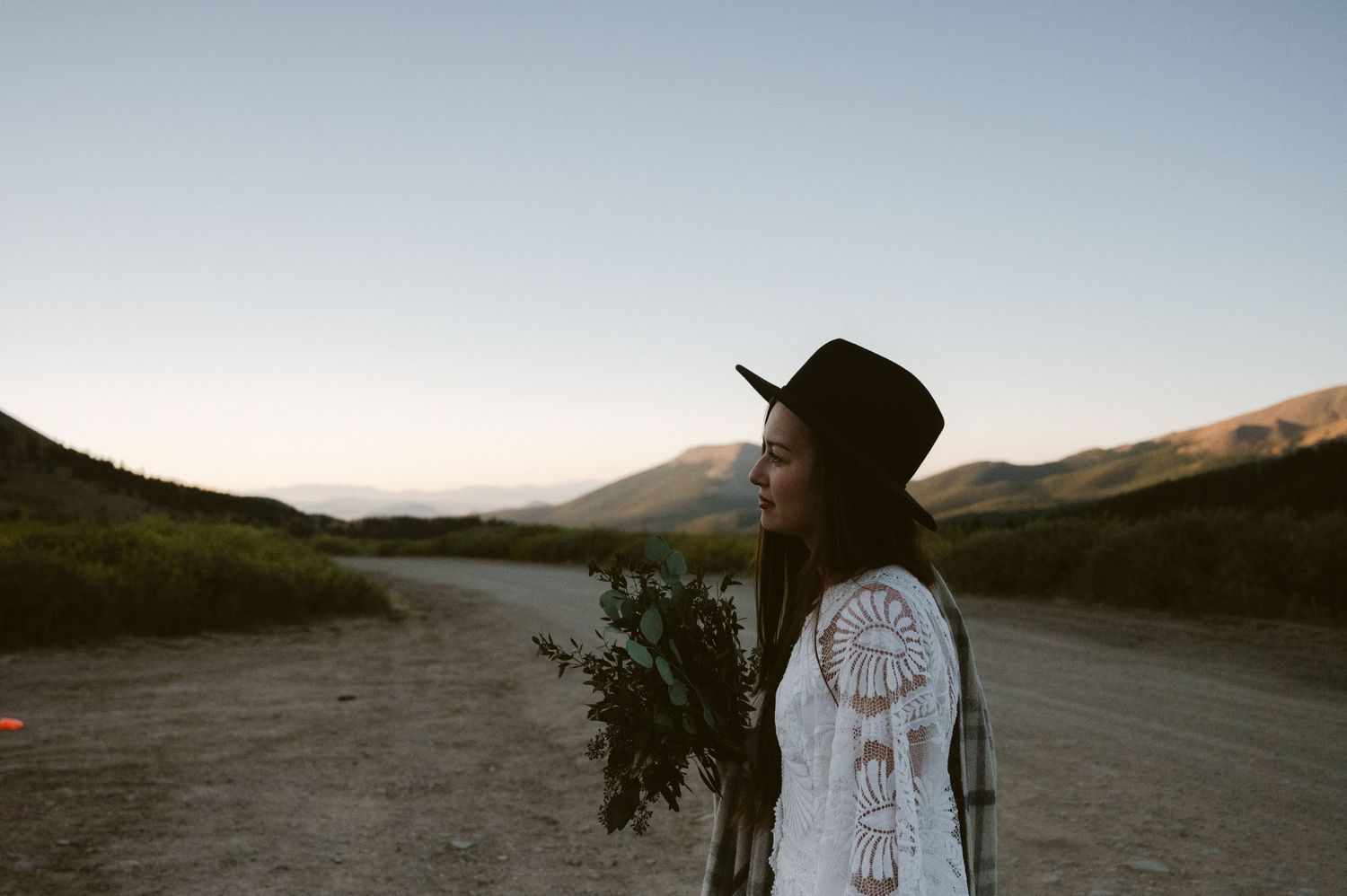 A person in a white dress and black hat stands on a dirt road with mountains in the distance.