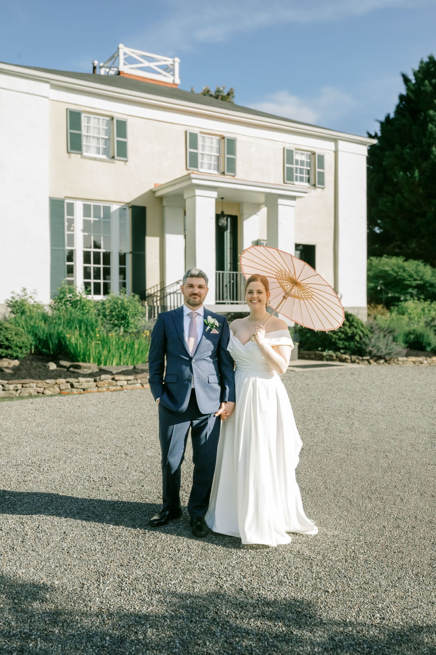 A couple stands in formal attire holding a peach parasol in front of a white colonial mansion.