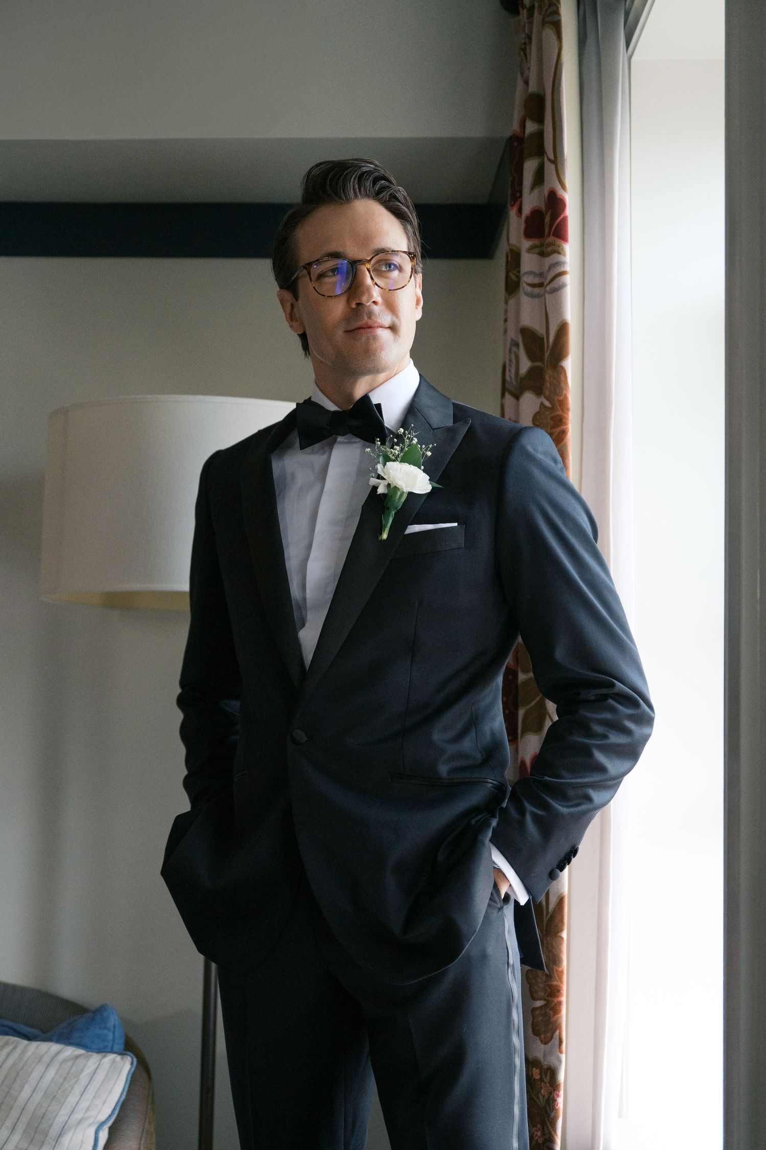 A groom in a classic black tuxedo with bow tie and boutonniere poses by a window.