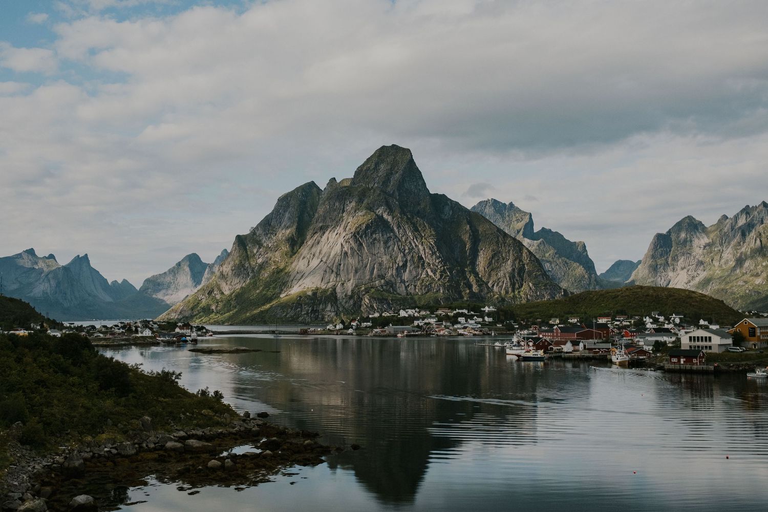 Dramatic mountain peaks reflect in calm fjord waters with small coastal village nestled along the shoreline in Norway.