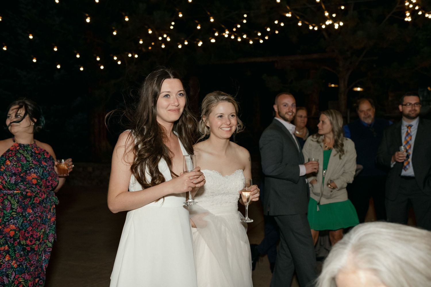 Wedding reception under string lights with guests mingling and holding drinks.