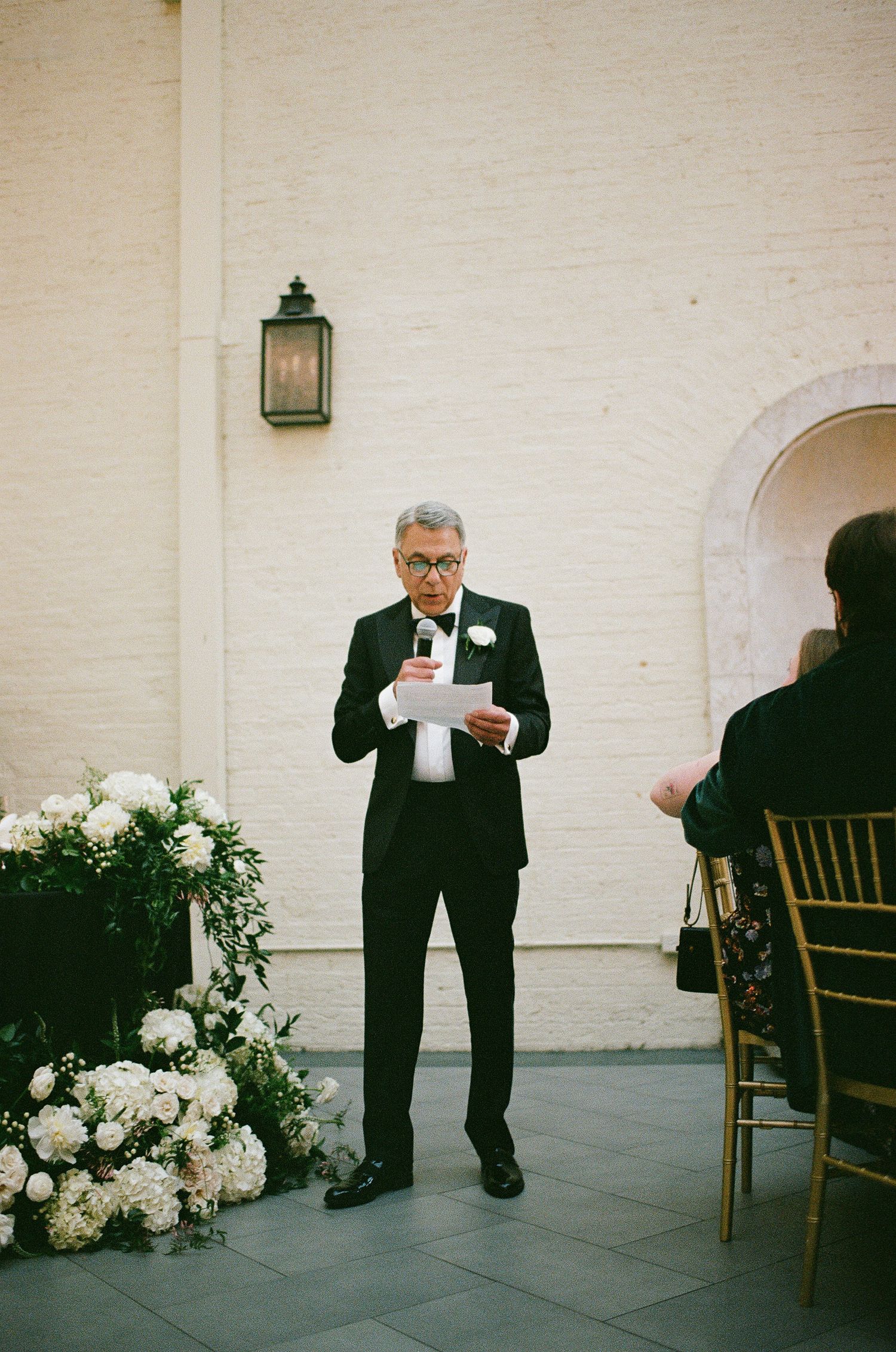 Wedding ceremony speaker in formal attire delivers remarks next to white floral arrangements.